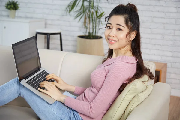 坐在沙发上的亚洲微笑的妇女和工作在膝上型电脑 — 图库照片