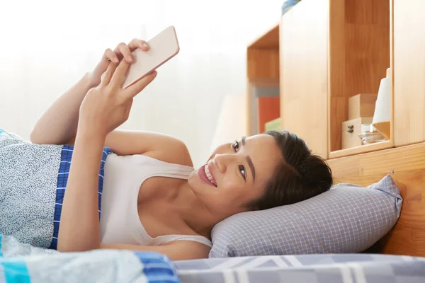 ベッドに横たわっていると 彼女の電話でアプリケーションを使用してかなりアジア女性を笑顔 — ストック写真