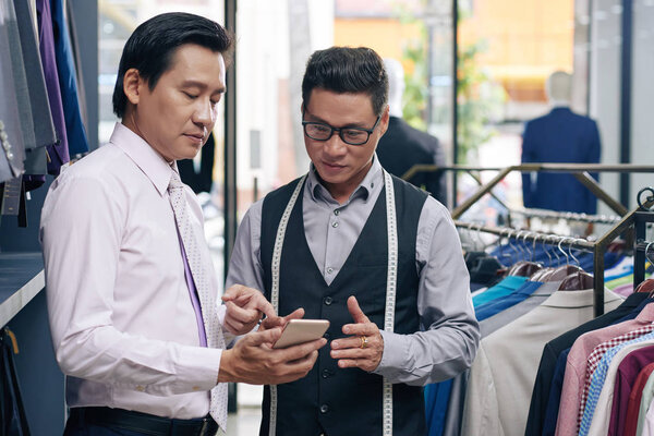 Зрелый азиат показывает фото костюма, который он хочет купить помощнику продавца
