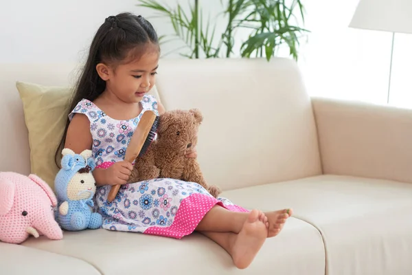 Kanepede oturan ve oyuncak ayısının fırçalama küçük kız