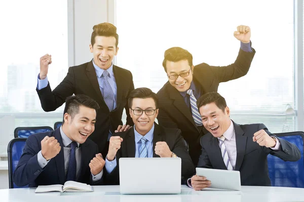 Cheerful Asian employees wearing classical suits gathered together in front of laptop and celebrating success