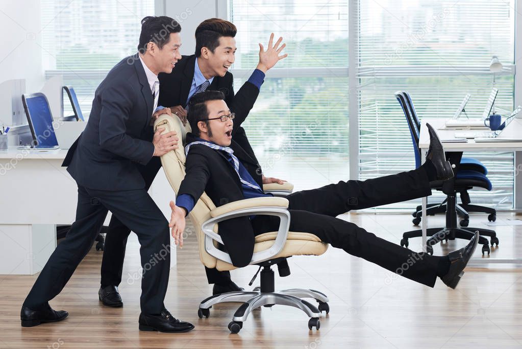 Asian white collar workers pushing swivel chair with colleague and laughing