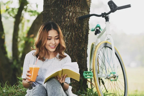 Gülümseyen güzel kadın içme güler yüzlü lezzetli ve ağacın altında bir kitap okuma