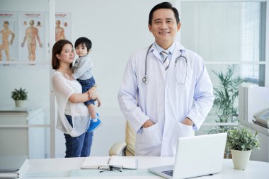 Asyalı doktor beyaz ceket kamera, anne ve oğlu arka plan üzerinde seyir 