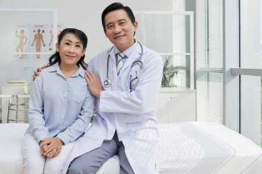 modern ward yatağın üzerinde otururken kameraya bakarak önemli hasta ve orta yaşlı Asya Doktor gülümseyen