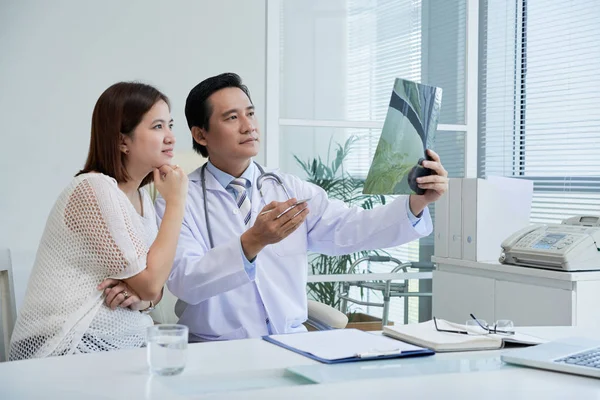 Selbstbewusster Chirurg Sitzt Schreibtisch Und Erklärt Hübschen Asiatischen Patienten Röntgenergebnisse — Stockfoto