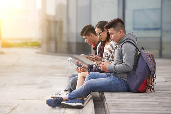 Group of Asian students sitting outdoors and surfing Internet on modern digital tablets