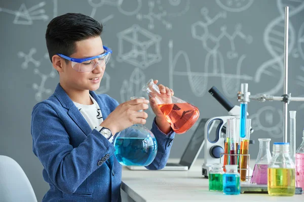 安全を身に着けている才能のある若手研究者は 化学教室で実験を遂行しながらフラスコに赤い液体を注ぐをゴーグルします — ストック写真