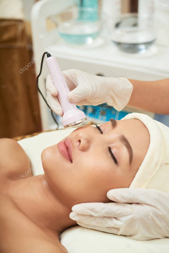 beautician hands making ultrasound facial procedure while pretty Asian client lying on treatment table with closed eyes