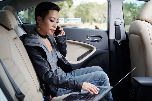 レザー ジャケットの車に座って 電話をかけることおよびラップトップに取り組んでの若い女性 — ストック写真