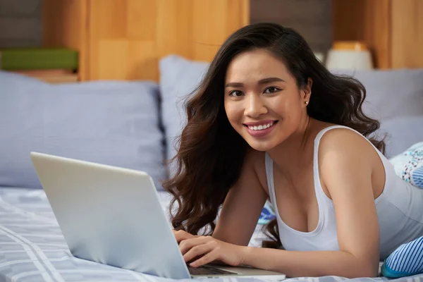 ベッドで横になっていると ノート パソコンでインターネットを閲覧しながら歯を見せて笑顔でカメラ目線陽気なアジア女性 — ストック写真