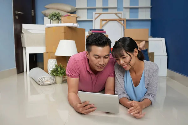 Asian couple using digital tablet and lying on floor in new flat with packed household stuff