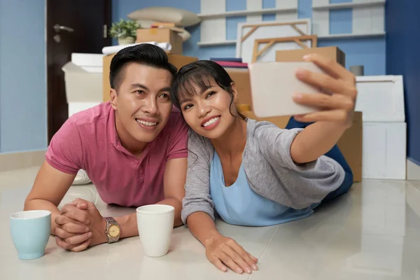 若いアジアのカップルのスマート フォン Selfie をとり パック ボックスと新しいアパートの床に横たわっている間幸せそうに笑って — ストック写真