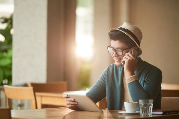 在数字平板电脑上阅读数据时戴着帽子打电话的年轻亚裔男子 — 图库照片