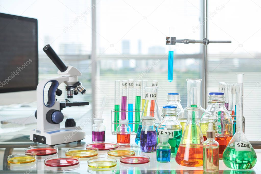 Interior of modern laboratory: desk with Petri dishes, test tubes, flasks and modern microscope on foreground, panoramic windows with blinds on background