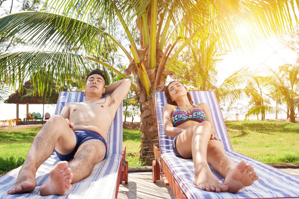 Young Vietnamese couple sunbathing on chaise-lounges under palm