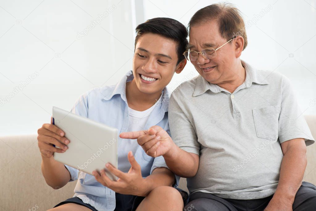Cheerful Asian teenager boy and his grandfather looking on tablet screen