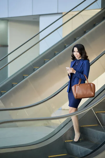 Pretty young asian woman in suit browsing smartphone while moving down escalator, full length