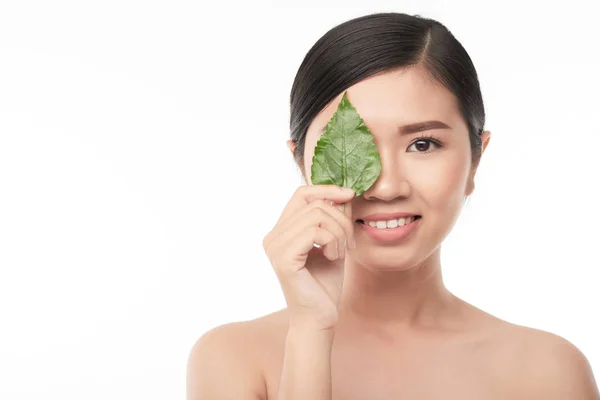 微笑的年轻亚洲妇女以无瑕疵的皮肤覆盖一只眼睛绿色叶子 — 图库照片