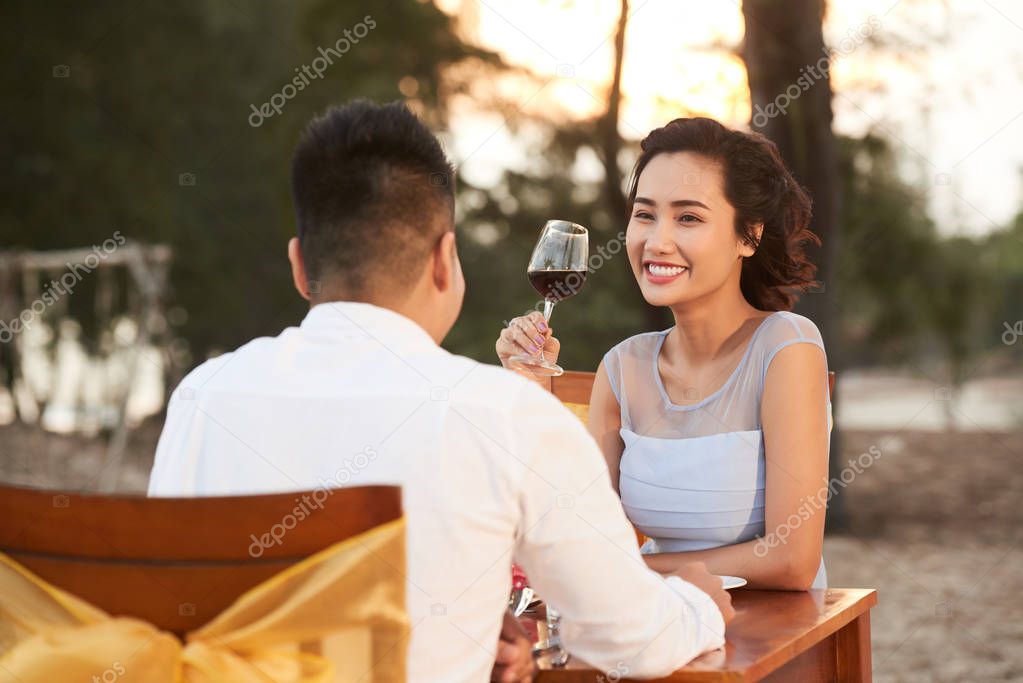 Attractive young woman with charming smile savoring red wine and listening to her loving boyfriend with interest while spending evening at beach restaurant