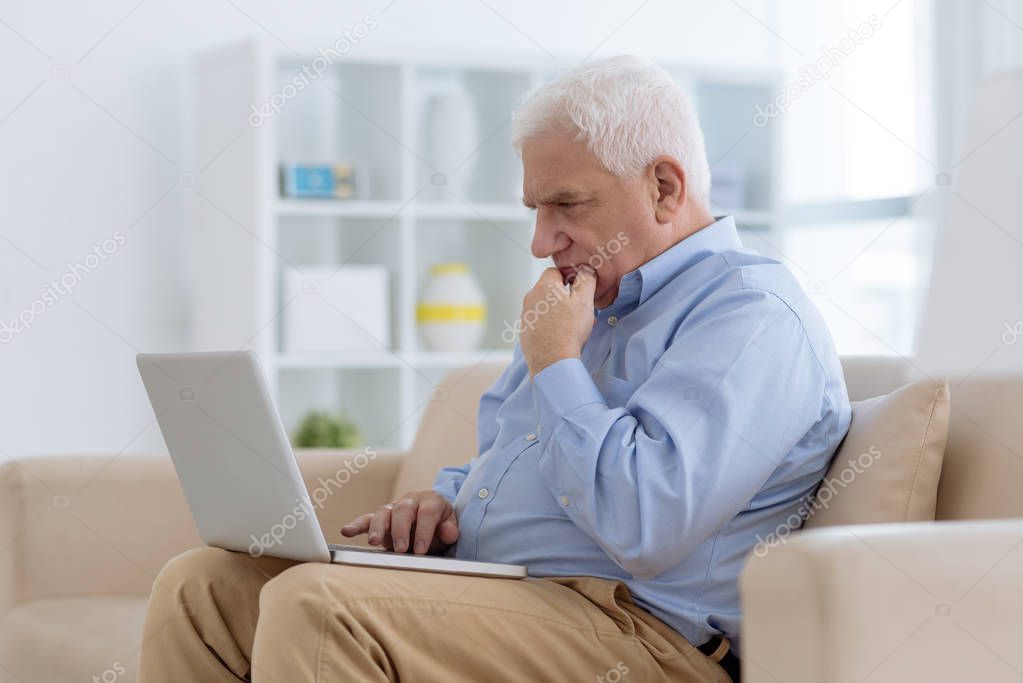 Pensive senior man sitting on sofa and working on laptop