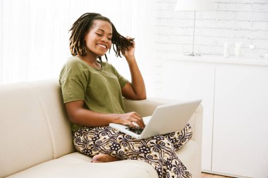 Dizüstü bilgisayar kullanarak ve ışık odada evde kanepede oturan oldukça neşeli kadın 