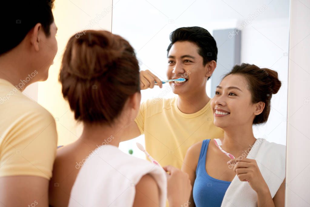 Cheerful young Asian couple looking at mirror and brushing teeth together