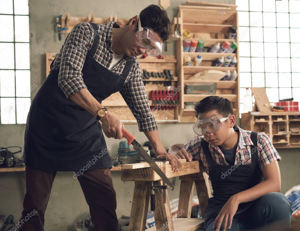 Vietnamese teenage boy looking how his father sawing wooden plank