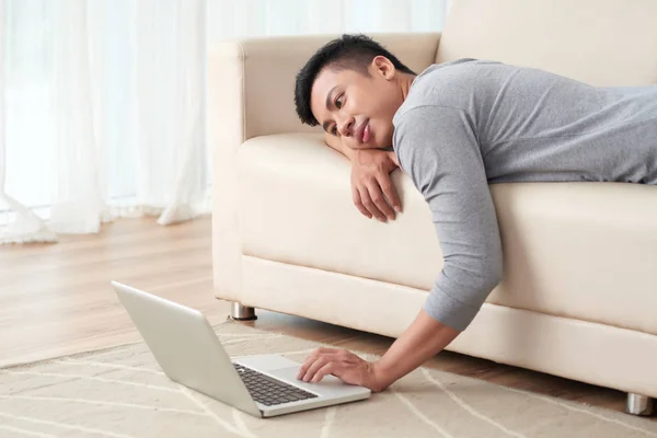 懒惰的亚洲人躺在沙发上 看着笔记本电脑上的东西 — 图库照片