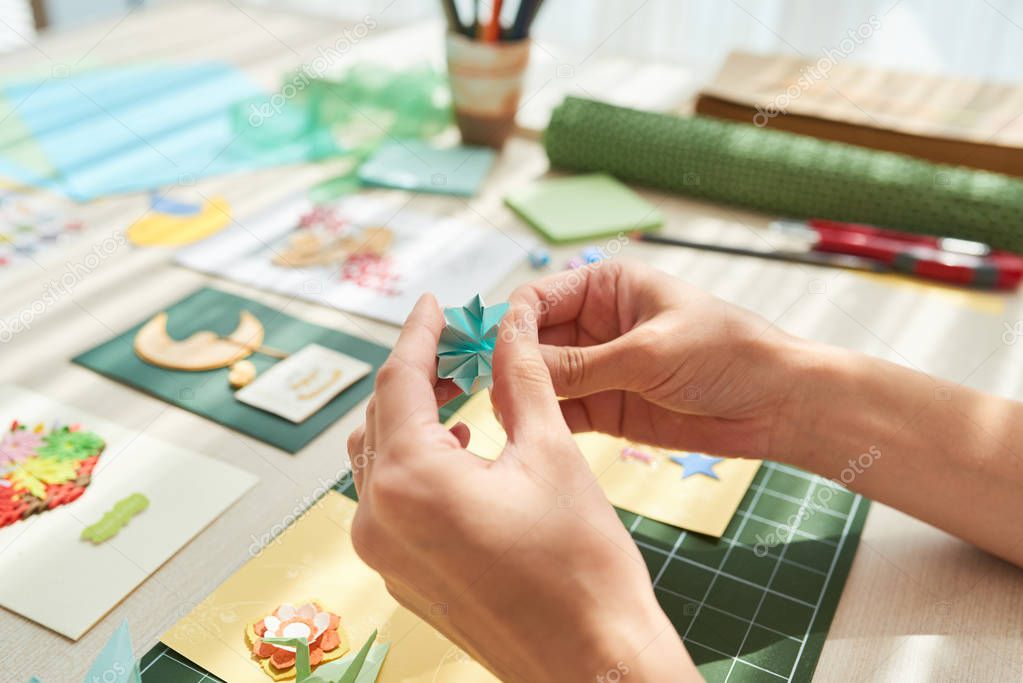 Unrecognizable young woman making decorative elements for greeting card while sitting at wooden table