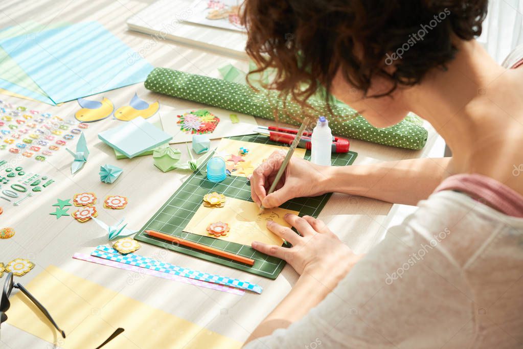 woman making colorful greeting card while sitting at wooden table, decorative items and tools lying on surface