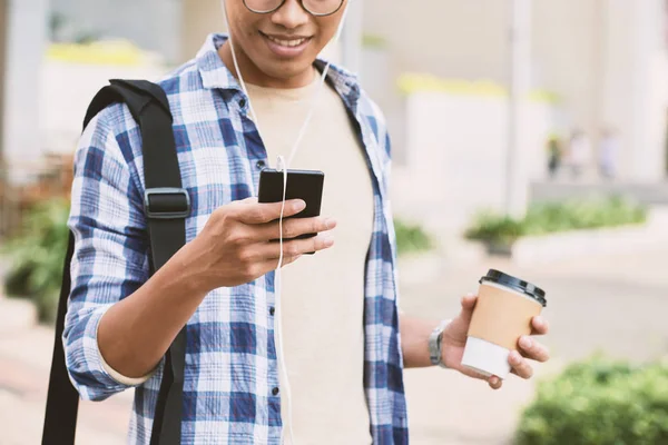 Przycięte Zdjęcie Uśmiechnięty Mężczyzna Azji Słuchawkach Przeglądania Smartfonem Trzymając Kubek — Zdjęcie stockowe