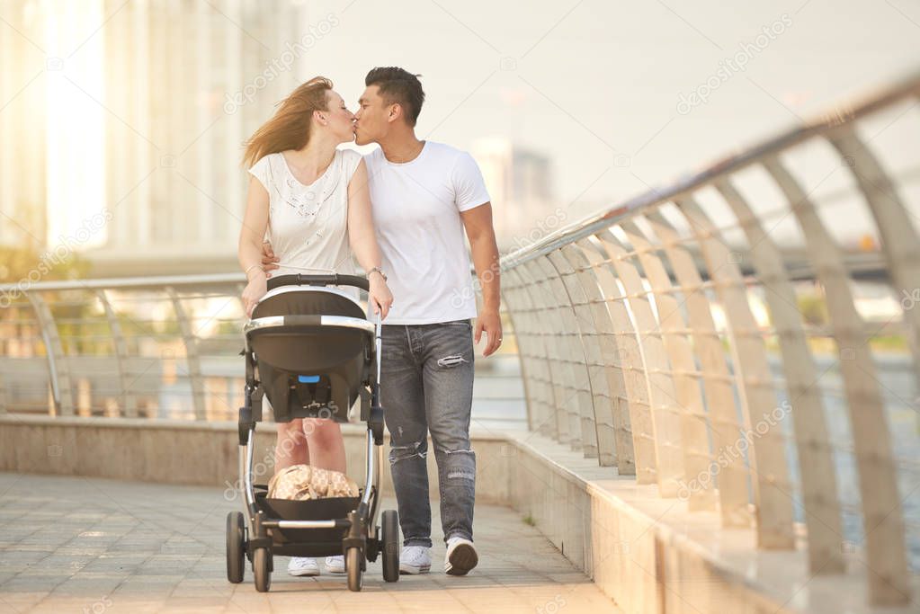 Attractive Caucasian woman kissing Asian husband while pushing baby carriage and walking along river 