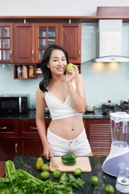 Genç Asyalı kadın yeşil elma tutarak ve taze meyve ve sebze tablo ile mutfakta ayakta beyaz top yerleştirmek