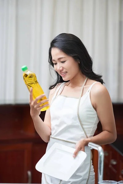 Asian woman in apron standing with dish near sink and exploring detergent bottle