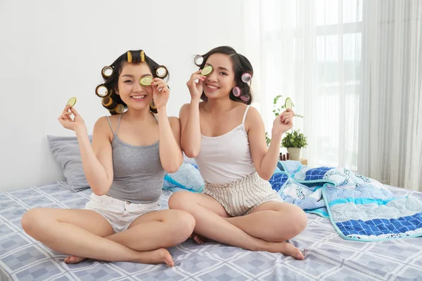 つの若いアジアの女性は笑みを浮かべて スタイリッシュな寝室で快適なベッドの上に座って顔近くキュウリのスライスを維持 — ストック写真