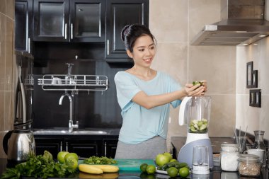 Çekici Asyalı kadın gülümseyerek ve taze malzemelerle blender smoothie mutfakta hazırlarken koyarak