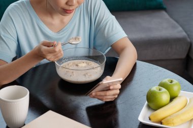 Anonim Bayan smartphone tarama ve müsli masada oturma odasında otururken yeme atış yukarıdan