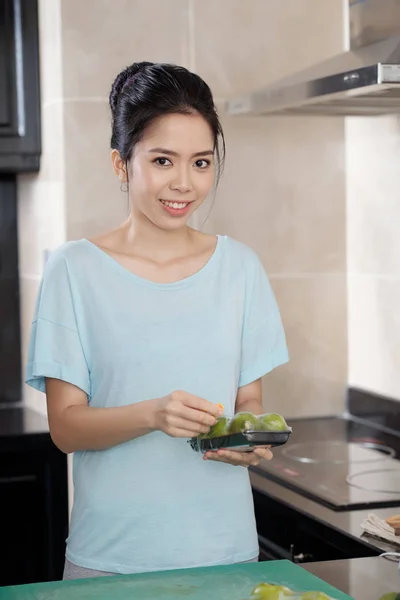 美しいアジアの女性笑顔と台所に立って 緑の果物のパックを保持しながらカメラ目線 — ストック写真