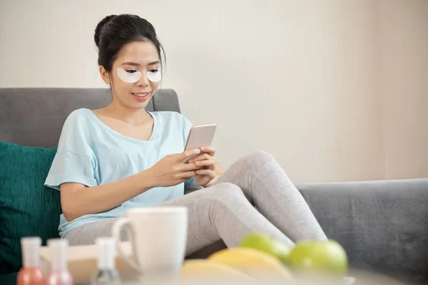 迷人的亚洲女性与眼睛不足的补丁微笑 并使用智能手机 而坐在舒适的沙发上在客厅 — 图库照片