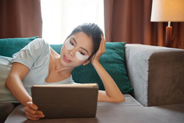 漂亮的亚洲女性与眼睛不足的补丁微笑和浏览平板电脑 而躺在舒适的沙发上 在客厅 — 图库照片