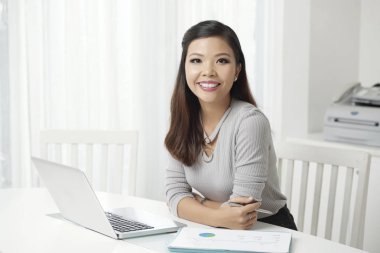 Kameraya gülümseyen ışık beyaz ofis dizüstü bilgisayar ile masada oturan genç modern etnik kadın