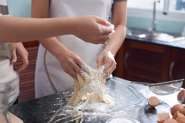 Crop shot of anonymous women wearing aprons kneading and sprinkling cookie dough with flour on kitchen table