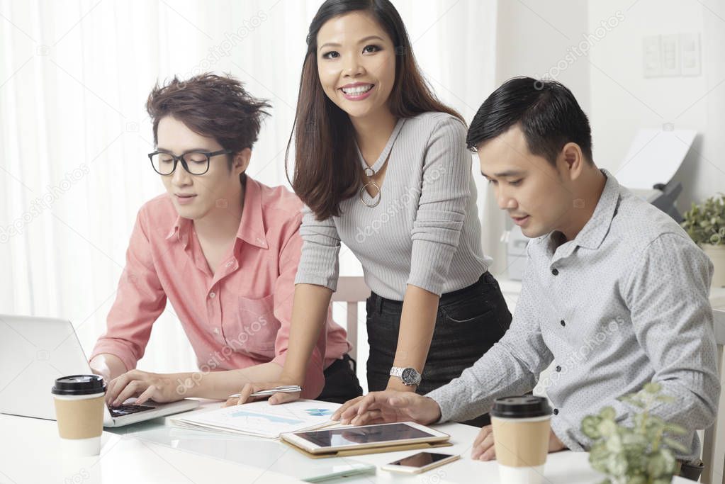 Cheerful Asian woman and met at desk in light office having coffee while working with gadgets