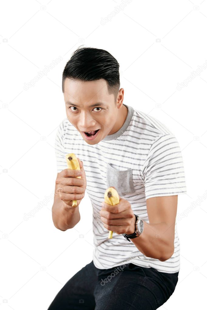 Portrait of young playful Asian man fooling with bananas and looking at camera while standing  against white background