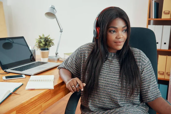 Modern African American woman in earphones sitting at table with laptop and papers in office