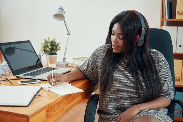 当代黑人妇女在办公室的桌旁工作和写作时听音乐 — 图库照片