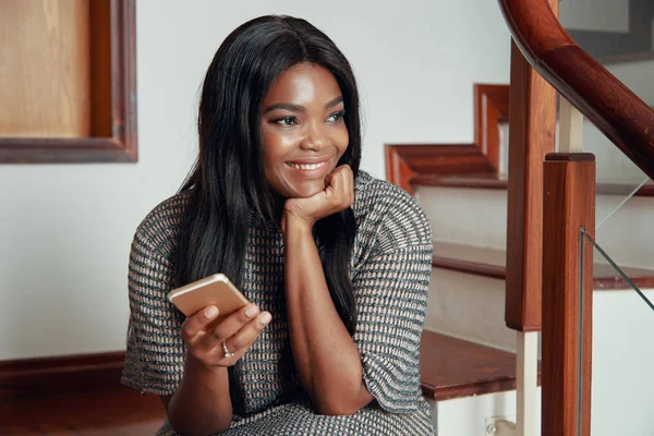 Adult smiling woman holding phone and sitting on wooden stairs of house looking away