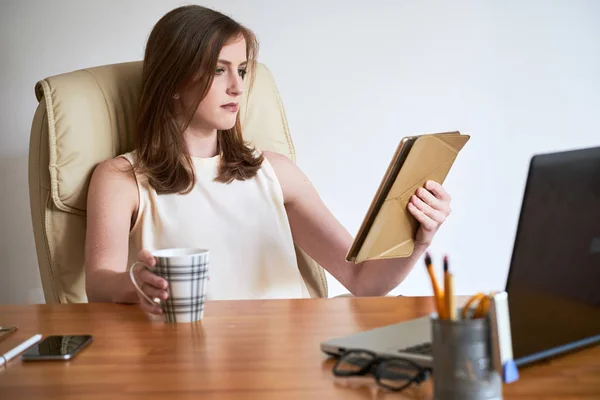 严肃的现代女性在办公桌上看平板电脑 一边喝咖啡 — 图库照片