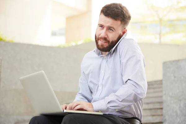 屋外のコンピューターで作業するときに 電話で話している陽気な笑みを浮かべて青年実業家 — ストック写真
